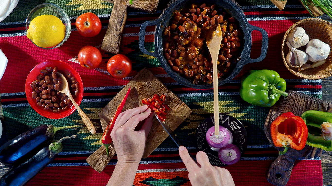 Et par hender som lager chili sin karne. Det er et bord full av mat med en gryte med chili sin carne i midten av bordet.